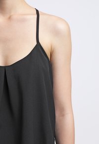Top negro sin mangas con tirantes finos. Presenta un diseño plisado en el escote y una textura de tela suave.