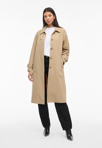 Donna in piedi che indossa un lungo trench beige, una maglietta bianca, pantaloni neri e stivali col tacco neri su uno sfondo bianco.