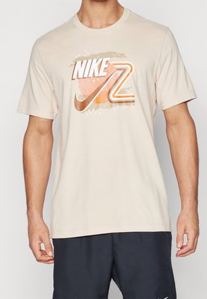 Camiseta beige de manga corta con logo y gráfico de Nike en naranja y blanco, llevada por una persona con los brazos relajados a los lados.