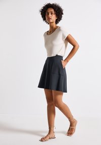 Haut à manches courtes beige associé à une jupe plissée noire avec des poches avant, complété par des sandales à lanières beige. Textures de tissu lisses.