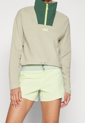 Chaqueta de forro polar beige y verde con cremallera hasta el cuarto y cintura con cordón, usada con pantalones cortos deportivos verde claro.