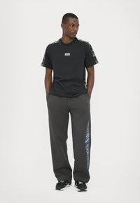 Black t-shirt with short sleeves and a logo; grey sweatpants with blue text on the side; model wearing black sneakers.