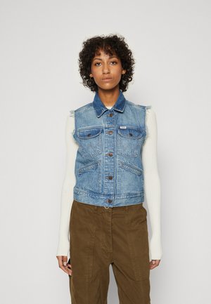 Gilet in denim azzurro chiaro con colletto, quattro tasche frontali e chiusura a bottoni. Indossato sopra una maglietta bianca a maniche lunghe e abbinato a pantaloni marroni.