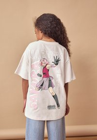 Šviesios spalvos per didelis marškinėlis su grafiniu atspaudu, vaizduojančiu personažą su rožiniais plaukais rožiniame ansamblyje, greta teksto "NARUTO" ir "SAKURA".