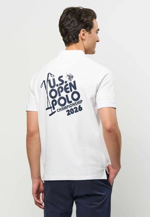 Mężczyzna w białym polo z tekstem „U.S. Open Polo Championship 2026” i grafiką pałki polo na plecach, stojący bokiem.