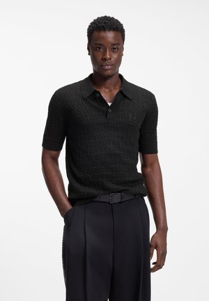 Mann trägt ein strukturiertes schwarzes Polohemd mit kurzen Ärmeln und hochtaillierte schwarze plissierte Hose mit einem schwarzen Gürtel, steht mit einer Hand in der Tasche.