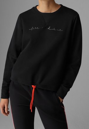 Personne portant un sweat-shirt noir avec le texte « fire and dice » en cursive et un pantalon noir noué avec un cordon de serrage orange vif.