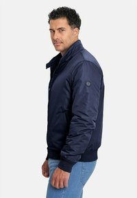 Chaqueta bomber azul marino con cuello alzado, textura acolchada, puños elásticos y cierre frontal con cremallera. Presenta un parche con logo en la manga.