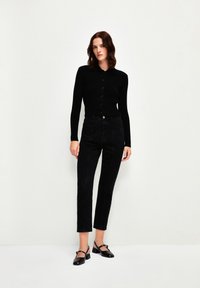 Cárdigan negro de botonadura con mangas largas, combinado con unos jeans negros de cintura alta y unos zapatos slingback negros con detalle de tira.