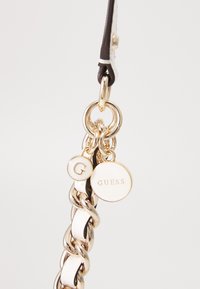 Metall-Schlüsselanhänger mit einer goldenen Kette, versehen mit einem weißen runden Anhänger mit der Aufschrift "GUESS" und einem kleineren runden Anhänger mit "G." Glatte und strukturierte Akzente.