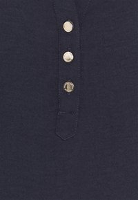 Mørkeblå henley skjorte med en glat tekstur, der har tre gyldne knapper, en strop og et lille broderet lomme-detail.
