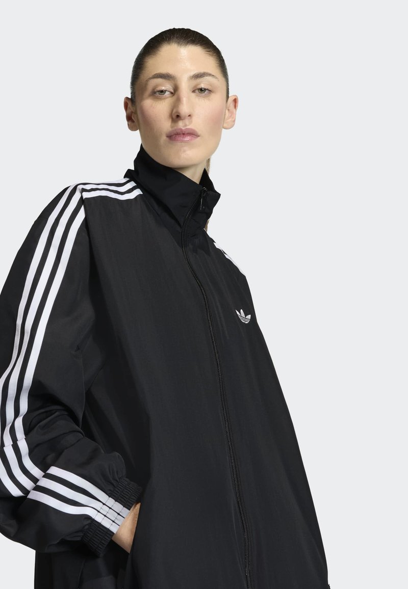Donna con i capelli legati indossa una giacca nera con zip, con strisce bianche Adidas sulle maniche e il logo Trefoil sul petto sinistro, con le mani in tasca.