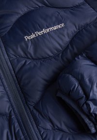 Marineblauwe pufferjack met een gequilt ontwerp, zichtbare stiksels en een Peak Performance-logo in zilver. Voorzien van een ritssluiting en een zachte textuur.