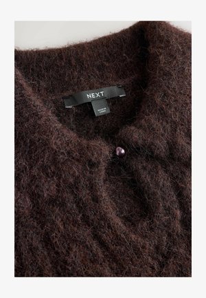 Cardigan en tricot marron et duveteux avec un col rond. Présente un bouton violet et une étiquette indiquant "NEXT", précisant la marque et l'origine.