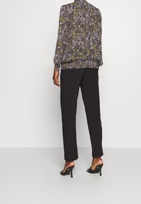 Blumenbluse mit langen Ärmeln, gerafften Manschetten und schwarzem Hintergrund, kombiniert mit schwarzen maßgeschneiderten Hosen und offenen, schwarzen Pumps.