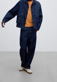 Modrá džínová bunda se dvěma předními kapsami, spárovaná s oranžovým tričkem a volnými modrými džínovými kalhotami. Světle zbarvené tenisky dotvářejí celý outfit.