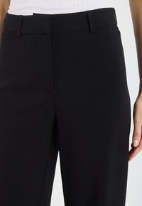 Pantalons noirs en tissu lisse et léger ; dotés d'un devant plat, d'une poche sur le côté et d'une taille ajustée.