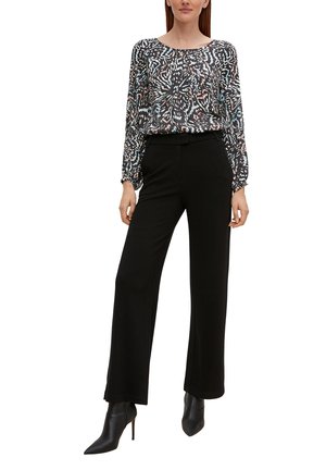 Vrouw met een blouse met lange mouwen en een meerkleurig patroon, zwarte broek met wijde pijpen en zwarte, puntige enkellaarzen met hoge hakken.