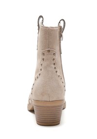 Botín de tobillo de ante beige con tachuelas a lo largo del tacón. Cremallera lateral y un tacón de madera apilada. Textura suave y forma redondeada.