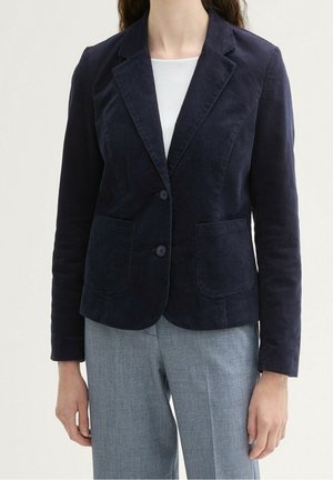 Frau trägt ein figurbetontes marineblaues Sakko mit zwei Knöpfen und aufgesetzten Taschen, kombiniert mit hellgrauen Hosen und einem weißen Oberteil darunter.