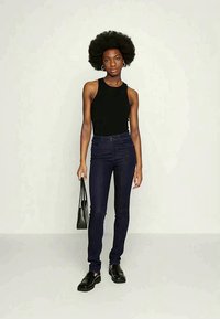 Moteris su afros stiliumi, vilkinti juodą be rankovių viršų, tamsias siauras kelnes, juodus loaferius ir laikančią juodą rankinę.