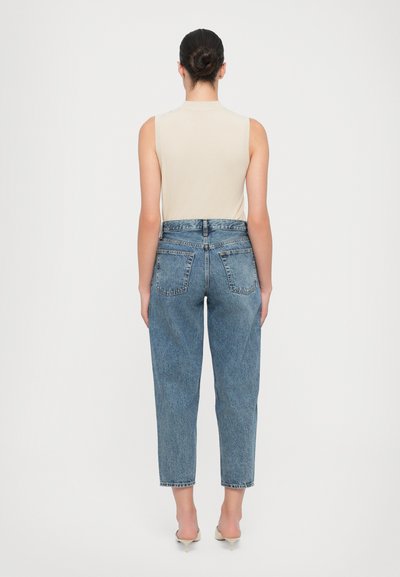 Beige mouwloze top gecombineerd met high-waisted blauwe denim jeans. De jeans hebben een relaxte pasvorm, vijf zakken en een onafgewerkte zoom.