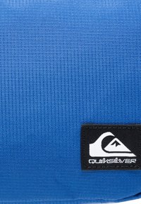 Sininen kangas, jossa on teksturoitu pinta, ja yhdellä puolella musta Quiksilver-logo-patch. Muoto on suorakaiteen muotoinen.