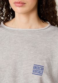 Siva majica iz bombažne mešanice z širokim izrezom in raznabljenim robom. Opozen modri logotip na sprednjem levem delu.