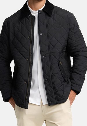 Mann trägt eine schwarze gesteppte Jacke über einem weißen Hemd mit Knöpfen und beigen Hosen, Hände in den Jackentaschen, vor einem einfarbigen Hintergrund.