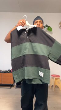 Czarna i zielona, pasiasta koszulka polo z długim rękawem na wieszaku, z białym kołnierzykiem, teksturowaną tkaniną i metką na dole.