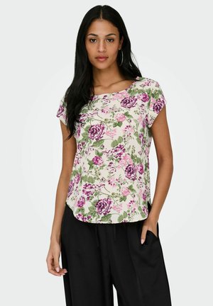 Woman wearing short-sleeve white blouse with pink and purple floral print, paired with loose black pants, standing with one hand in pocket.