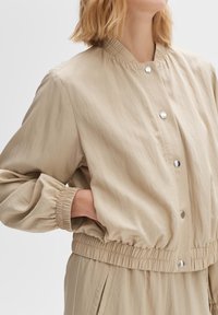 Beige bomber jacket with a ribbed collar, elastic waistband, and puffed sleeves. Features silver snap buttons along the front.