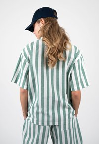 Femme aux longs cheveux ondulés portant une chemise et un pantalon à rayures verticales vertes et blanches, face tournée, casquette bleu marine portée à l’envers.
