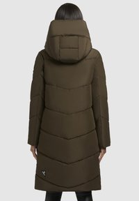 Lang, olijfgroene pufferjas met chevronstiksels, een dikke capuchon en een zwart logoaccent. Het materiaal lijkt zacht en geïsoleerd.