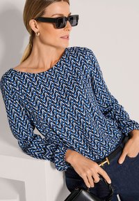 Blue patterned blouse featuring geometric shapes, long sleeves with elastic cuffs, paired with a black belt and a small black handbag.