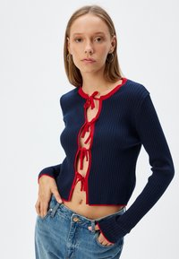 Cardigan a coste navy con accenti rossi, caratterizzato da maniche lunghe e chiusura anteriore con nodo. Lunghezza media, texture morbida e design aderente.