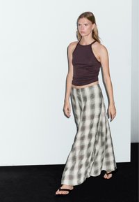 Top senza maniche marrone abbinato a una gonna lunga fluida a scacchi bianco e nero. Modella che indossa sandali piatti, su uno sfondo minimalista.