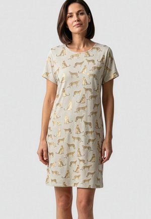Vrouw draagt een beige korte mouw jurk met gouden metallic jachtluipaardprint, staand tegen een effen lichte achtergrond.