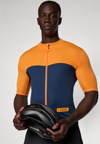 Maillot de cyclisme à manches courtes en orange et bleu marine, avec une fermeture éclair mi-hauteur à l'avant et une étiquette "LE COL". Le modèle tient un casque de cyclisme noir.
