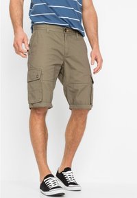 Pantaloni cargo color sabbia con tasche laterali, risvolti e chiusura a bottone, abbinati a sneakers in bianco e nero e una camicia a righe blu.