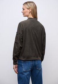 Mujer de cabello rubio con una chaqueta bomber de canalé en color oliva oscuro y pantalones vaqueros azules, de pie frente a un fondo liso de color gris claro.