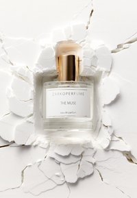 Flacon de parfum en verre transparent avec un capuchon doré, portant une étiquette blanche sur laquelle est inscrit "ZARKOPERFUME THE MUSE eau de parfum," sur fond de texture blanche craquelée.