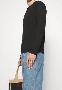 Person, der ein schwarzes Langarmshirt und eine hellblaue Jeans trägt, hält eine beige und schwarze Tragetasche mit roter Schrift.