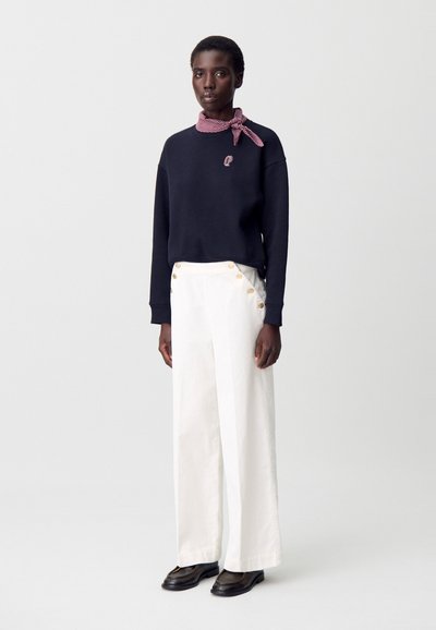 Sudadera de marina con logo, pantalones de pierna ancha blancos con botones dorados y una bufanda de cuadros rojos. Unos zapatos de cuero negros completan el look.