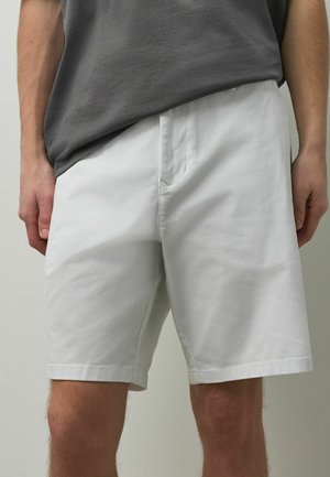 Mann trägt schlichte weiße knielange Shorts und ein lockeres graues T-Shirt, steht mit entspannten Händen an den Seiten.