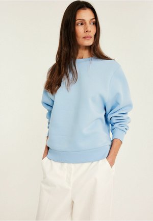 Kvinde med langt brunt hår iført en lyseblå sweatshirt og hvide bukser, stående med hænderne i lommerne og kigger til siden.
