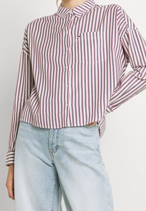 Vrouw die een rood-wit-blauw verticaal gestreept hemd draagt dat lichtjes in een lichtblauwe hoge taille jeans is gestopt.