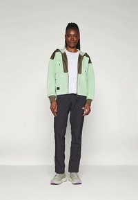 Γυναίκα που στέκεται φορώντας ένα φωτεινό πράσινο και καφέ φούτερ με φερμουάρ, λευκό μπλουζάκι, μαύρο παντελόνι και ανοιχτό γκρι αθλητικά παπούτσια με ανοιχτό μωβ σόλες.