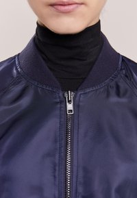 Chaqueta bomber de color marino con cierre de cremallera y cuello de canalé. Tejido liso y brillante con detalles de textura sutil. Suéter negro de cuello alto debajo.
