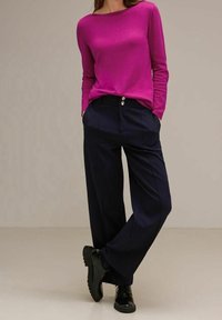 Kobieta ubrana w jasnoróżową bluzkę z długim rękawem, granatowe spodnie z szerokimi nogawkami i podwójnymi guzikami, oraz czarne sznurowane buty, stojąca z rękami w kieszeniach.